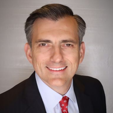 Professional headshot of smiling man in dark suit, light blue shirt, and red patterned tie
