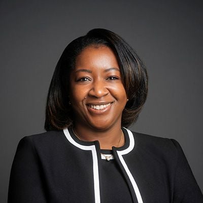 Professional headshot of a smiling woman in a black blazer with white trim against a gray background