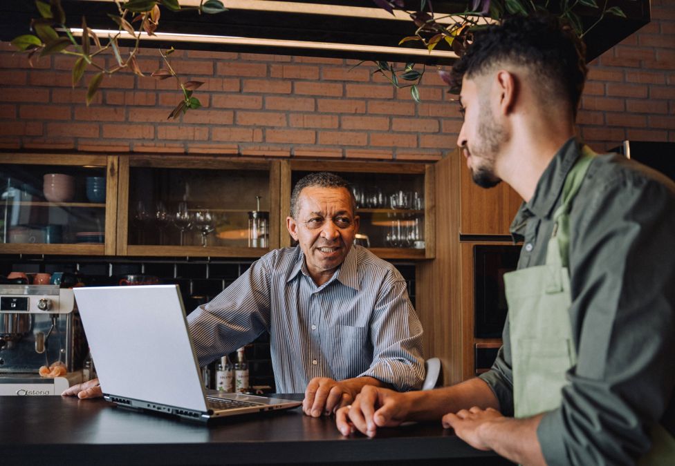 Two men converse over a laptop at the counter of a small café or restaurant.