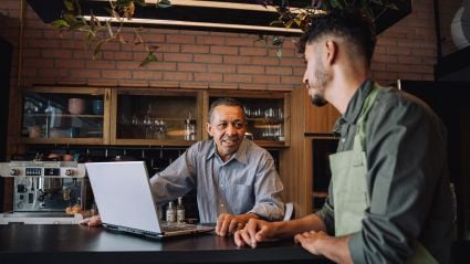 Two men converse over a laptop at the counter of a small café or restaurant.