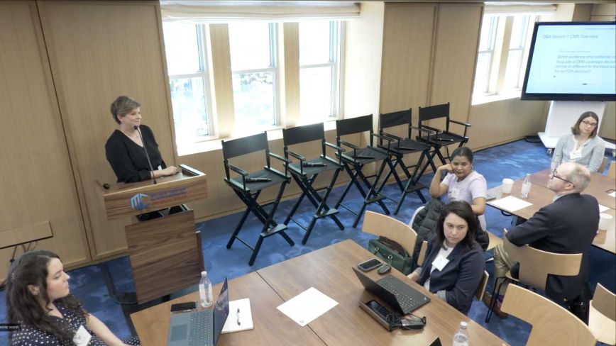 A high-angle view of a conference room during a Q&A session. A woman in a black top stands at a wooden podium on the left.
