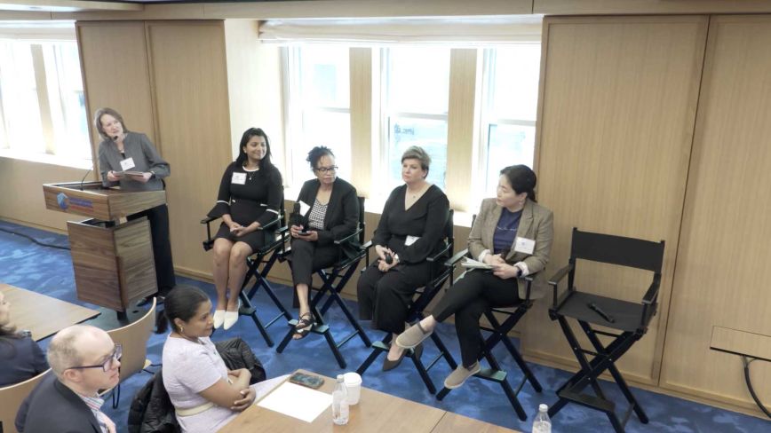 A diverse panel of four women seated in director's chairs during a professional workshop, with a moderator standing at a lectern and an audience in the foreground.
