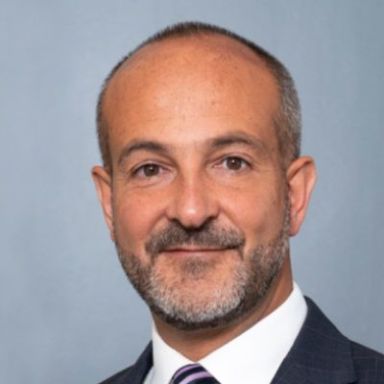 Professional headshot of smiling man with gray beard wearing dark suit and tie