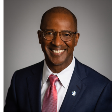 Professional headshot of a smiling man in a navy suit with pink tie and glasses