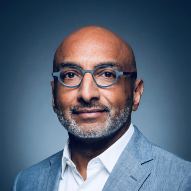Professional headshot of a smiling bald man with glasses wearing a light blue blazer